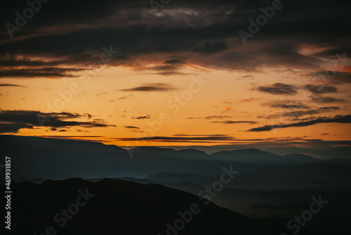 Scenic dawn mountain landscape with golden low clouds in valley among dark mountains silhouettes under sunset or sunrise sky. Vivid scenery with low clouds in mountain valley in illuminating color.