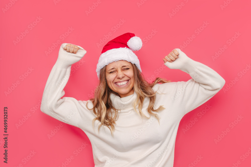 A young charming smiling blonde girl in a white warm sweater and a red Santa Claus hat is happy with her hands up at the advent of Christmas and New Year isolated on a color pink background A young charming smiling blonde girl in a white warm sweater and a red Santa Claus hat is happy with her hands up at the advent of Christmas and New Year isolated on a color pink background