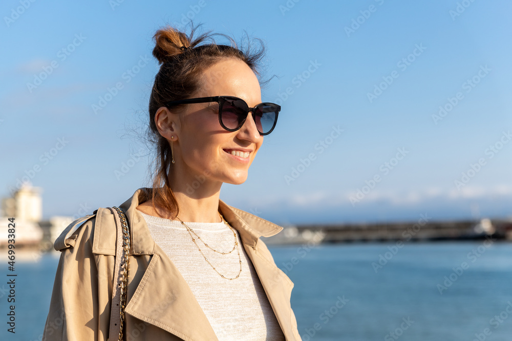 Yonug adult trendy stylish beautiful caucasian happy smiling woman enjoy walking by Yalta sea embankment on warm sunny day. Female person portrait wear jeans biege trench coat on urban city street