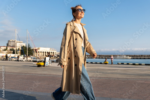 Yonug adult trendy stylish beautiful caucasian happy smiling woman enjoy walking by Yalta sea embankment on warm sunny day. Female person portrait wear jeans biege trench coat on urban city street photo