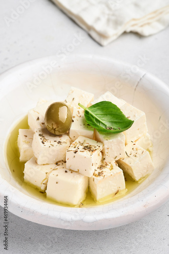 Feta cheese cubes with rosemary, olives and olive oil sauce in white bowl on light gray background. Traditional Greek homemade cheese. Selective focus.