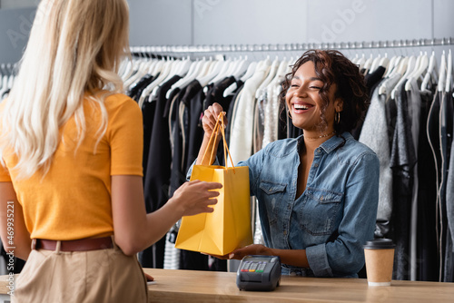 happy african american saleswoman giving yellow paper bag to client in boutique photo