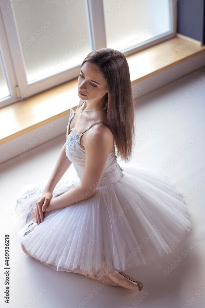 Young long haired ballerina dressed in white tutu costume pose in sit position on floor at ballet studio in natural ambient daylight atmosphere Young long haired ballerina dressed in white tutu costume pose in sit position on floor at ballet studio in natural ambient daylight atmosphere