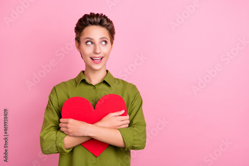 Photo of sweet excited young lady wear green blouse embracing big red heart looking empty space smiling isolated pink color background
