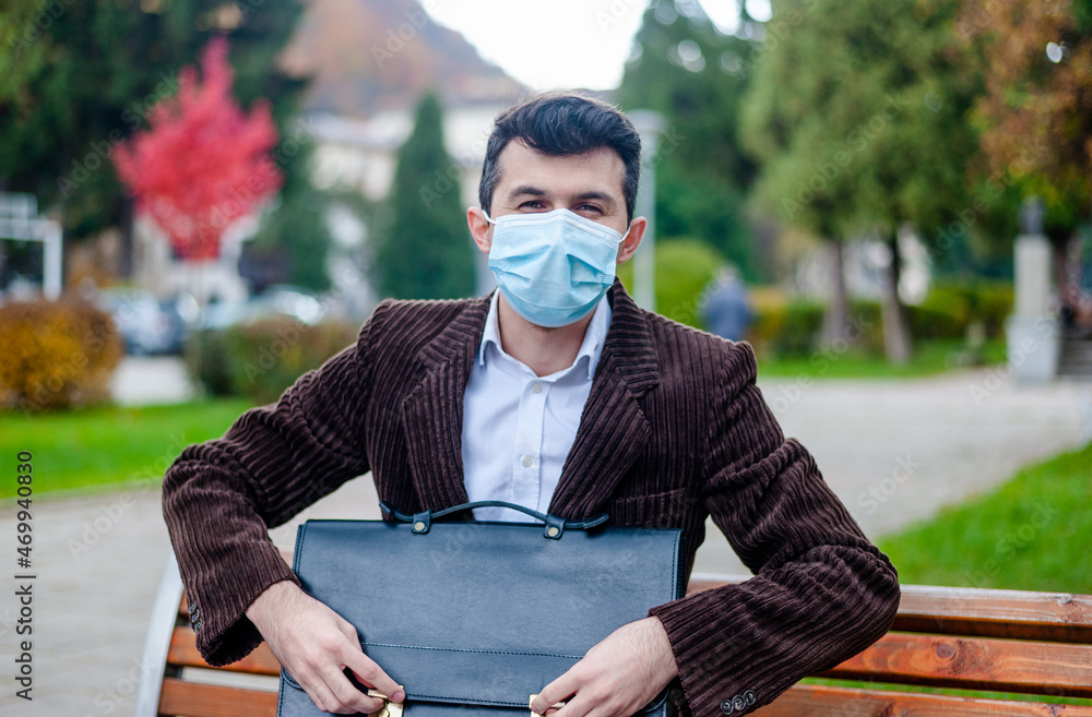 Fototapeta premium A happy businessman with a protective mask and briefcase sits on a park bench during a business break.