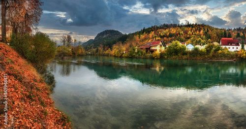 Panoramic amazing autumn landscape of a village in the Alps. View of the reflection of the fall colorful forest in the water of the river and of the mountain range in the distanceat sunset.