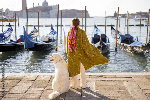 Young woman walking with dog near water canal with gondolas at saint Marks square in Venice. Woman wearing coat and colorful shawl in italian style. Maremmano abruzzese Italian sheepdog