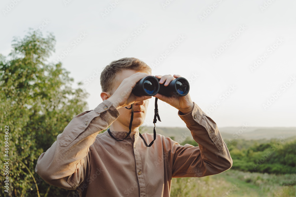 A man with modern binoculars against the sky and green forest.The concept of hunting, travel and outdoor recreation. Banner with copy space.A traveler or hunter is observing through binoculars. A man with modern binoculars against the sky and green forest.The concept of hunting, travel and outdoor recreation. Banner with copy space.A traveler or hunter is observing through binoculars.