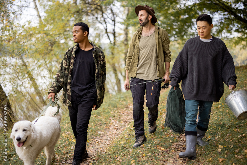 Multiracial male friends walking with Maremmano-Abruzzese Sheepdog and fishing equipment in nature. Concept of weekend and vacation in nature. Idea of friendship. Men between forest and river