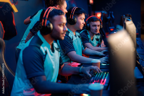 Group of multiethnic cybersport players in headsets sitting in front of computers and playing video games online indoors