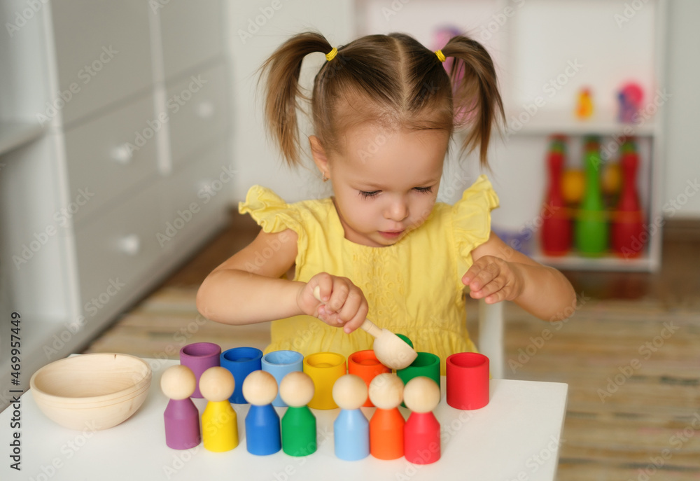 Preschooler kid learns colors and shapes by playing a wooden educational multicolored toy with balls and cups. A focused Child is sitting at a table in the children's room. Two - year - old girl Preschooler kid learns colors and shapes by playing a wooden educational multicolored toy with balls and cups. A focused Child is sitting at a table in the children's room. Two - year - old girl