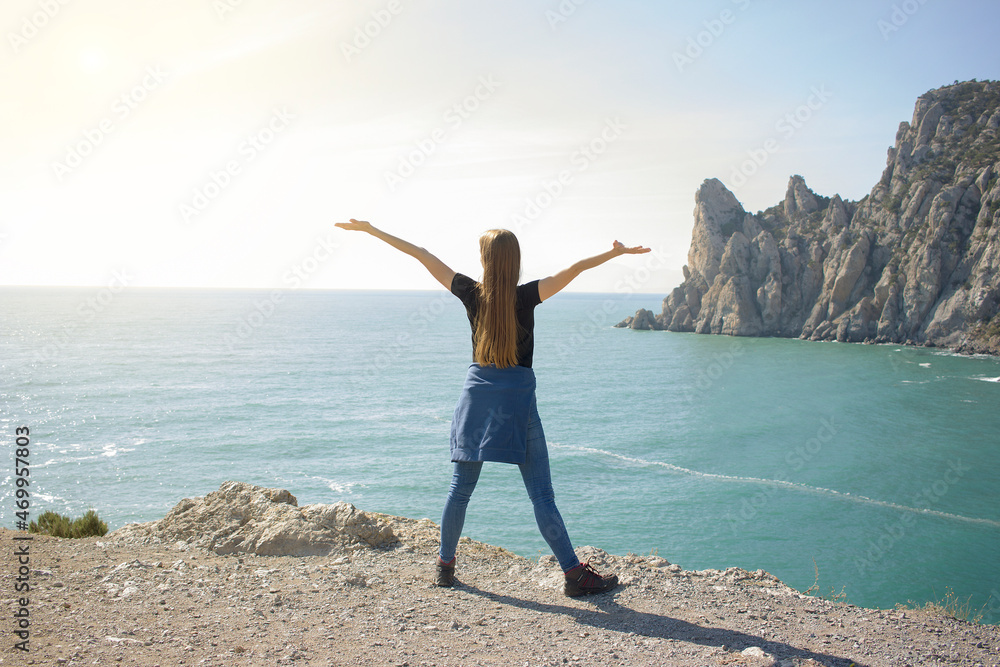 A girl stands on the edge of a cliff with her arms outstretched and looks at the sea and rocks. Sea travel concept. Admiration for nature.