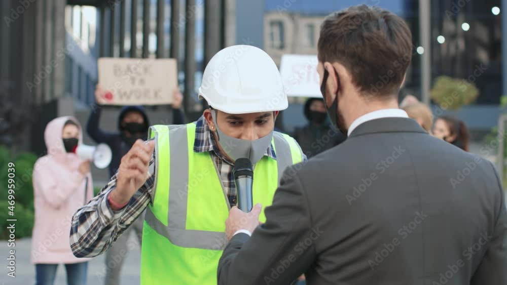 Medium closeup of builder in protective mask wearing helmet giving interview to male journalist standing outdoors in city centre around the protesting people Medium closeup of builder in protective mask wearing helmet giving interview to male journalist standing outdoors in city centre around the protesting people