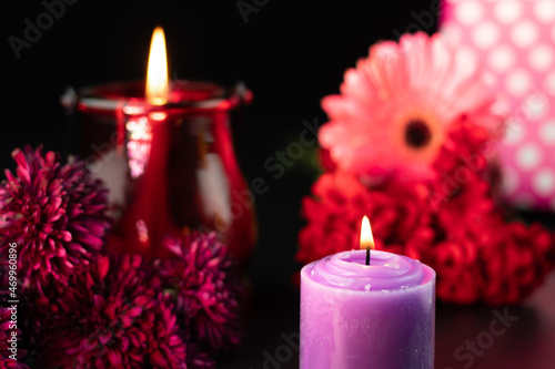 Flames Glowing From Designer Purple Or Lilac Pillar Candle With Colorful Fresh Floral Theme On Dark Black Background