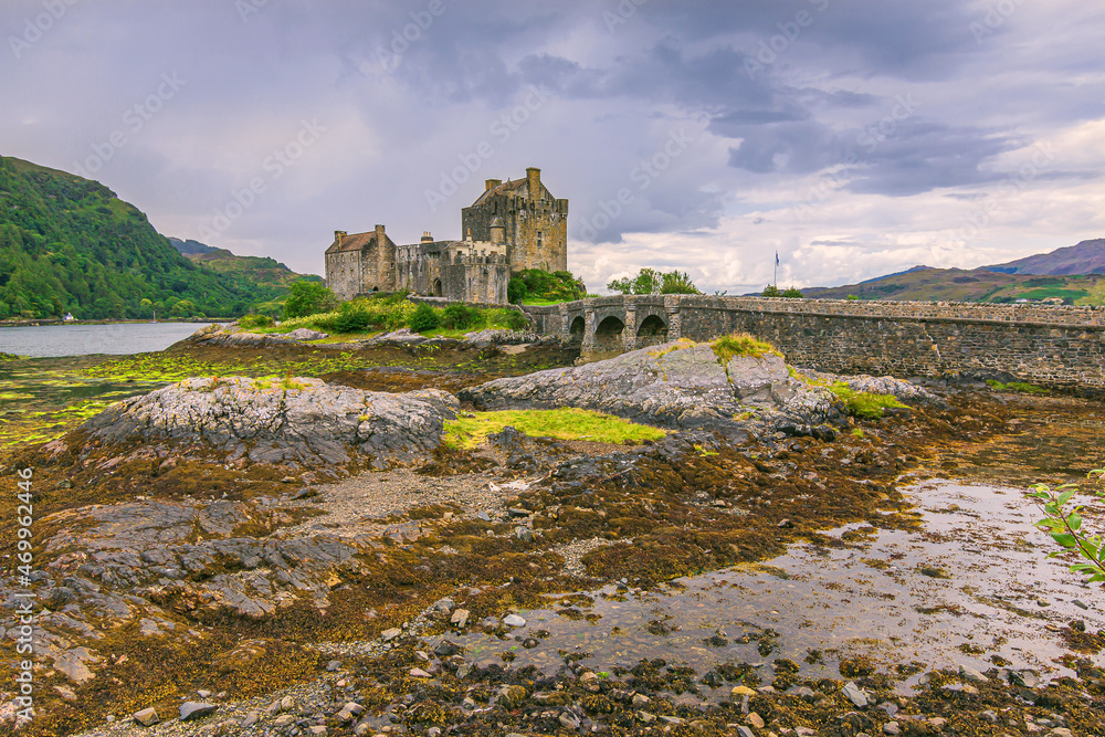 Historic building Eilean Donan Castle in Scotland in the daytime when the water is low with a stone bridge for pedestrians. Stones with algae in the foreground at low tide. Dark clouds in the sky