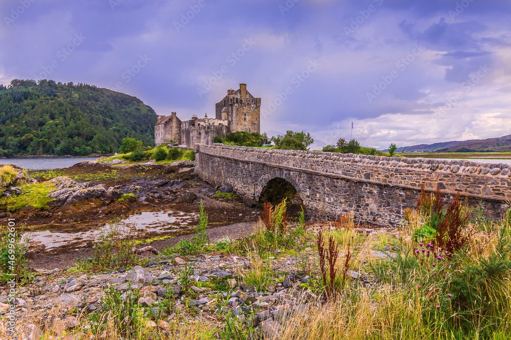 Eilean Donan Castle in Scotland with the historic stone bridge for pedestrians during the day at low tide. Hill and lake in the background from the building. Bushes, grass and stones in the foreground