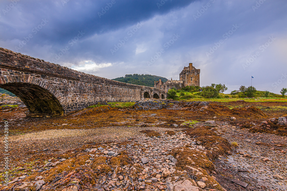 Historic castle in Scotland. Dry river bed under archway of the stone footbridge to Eilean Donan Castle. Island with the castle at low tide. Stones and grass in the foreground