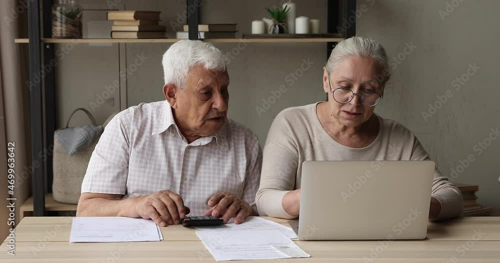 Focused mature spouses on pension sit at desk do domestic paperwork read check documents calculate pay utility bills on laptop online. Serious older couple review investment plan family budget economy Focused mature spouses on pension sit at desk do domestic paperwork read check documents calculate pay utility bills on laptop online. Serious older couple review investment plan family budget economy