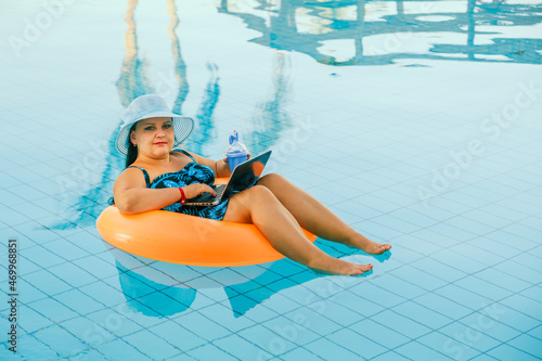 brunette woman in a sun hat and a swimsuit in the pool in a swimming circle works with a laptop.