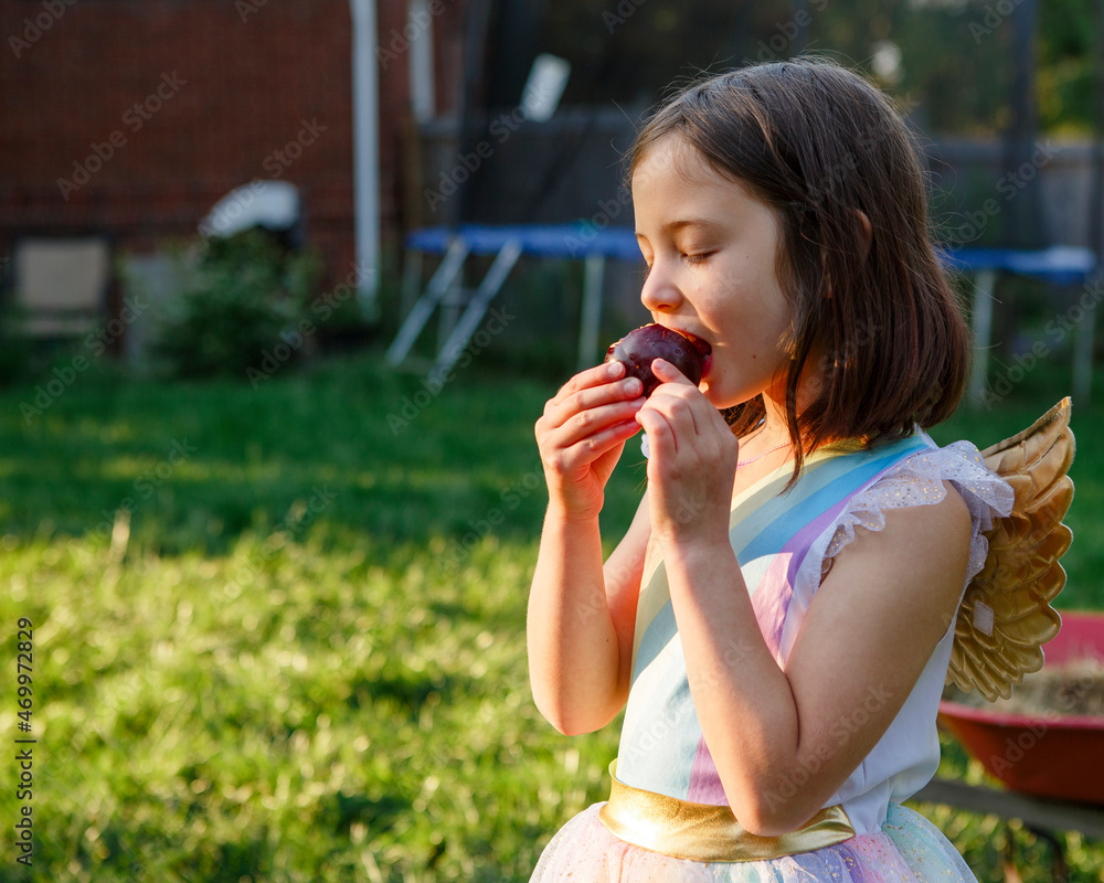 A small child in costume with golden wings eats juicy plum in sunshine A small child in costume with golden wings eats juicy plum in sunshine