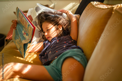 A child lays on couch in dappled window light reading chapter book photo