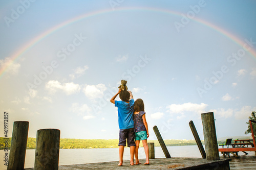 two children stand on deck under rainbow by sunlit lake with toy bear photo