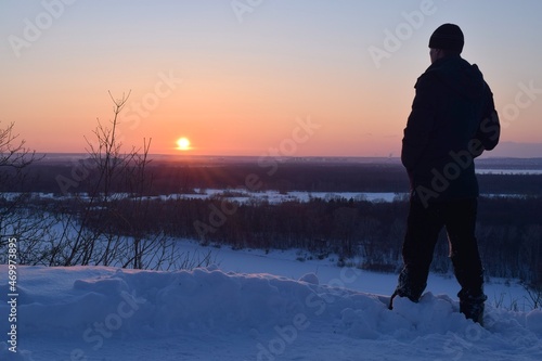 Silhouette of a man standing on a cliff by the river and looking at the winter sunset