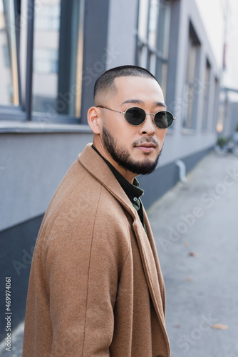 bearded asian man in autumn coat and sunglasses looking at camera near blurred building © LIGHTFIELD STUDIOS