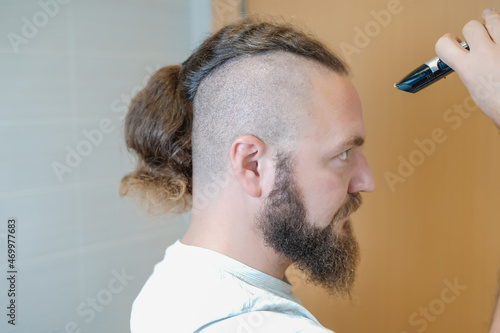 Caucasian man in profile with a beard and ponytail before cutting his hair with an electric trimmer.