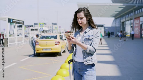 Young woman tourist brunette with long loose hair types on smartphone standing near suitcase by asphalt road waiting for taxi under bright summer sunlight. Taxi Hailing app concept