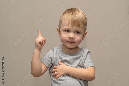 Cute boy in gray t-shirt blond European German age 3 years isolated on gray background shows, points a finger and communicates for advertisement layout, close-up layout with selective focus