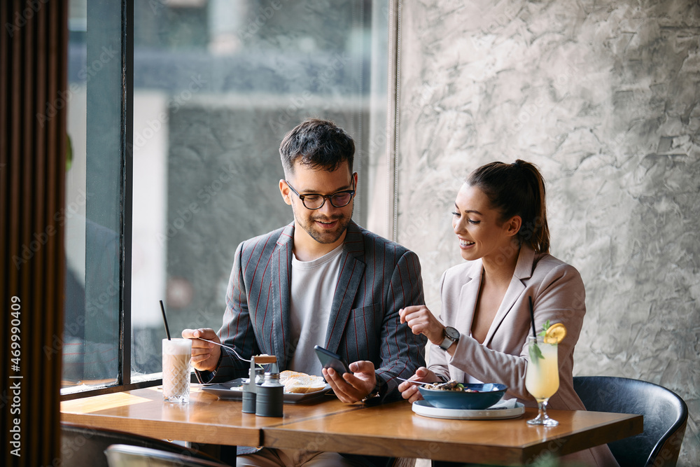 Obraz premium Young happy colleagues use mobile phone while having breakfast in cafe.