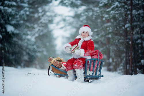 Little funny child dressed in Santa Claus red costume bringing presents in winter snowy forest. Christmas Eve.