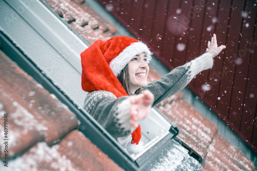 Young woman in a red and white Christmas hat looking out of a skylight window and smiling, totally excited about first snow. 