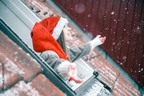 Bilde på lerret Young woman in a red and white Christmas hat looking up out of a skylight window and smiling, excited about first snow