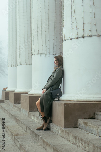 Thoughtful beautiful young woman in green coat standing near white columns. Girl in city