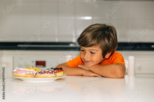 Blond boy sitting in the kitchen in front of four colorful donuts