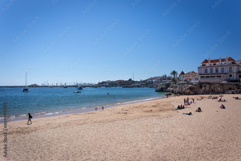  Beautiful Cascais Beach with people in the sand and beach houses and white boats in the distance over the beautiful blue sky and blue sea on a sunny spring day. 