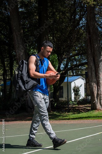 Man wearing a blue sleeveless T-shirt walking with a basketball ball and looking his phone in a basketball court