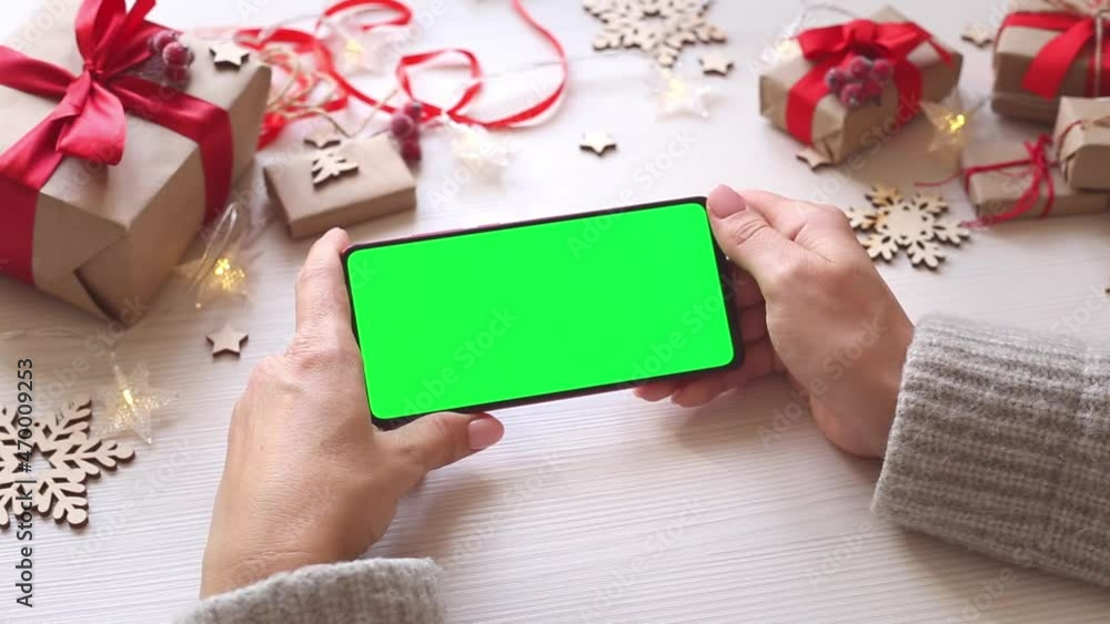 Woman holding smartphone with horizontal green screen on Christmas background with gifts. Mobile Phone with Chroma Key. Close up hands. Woman holding smartphone with horizontal green screen on Christmas background with gifts. Mobile Phone with Chroma Key. Close up hands.