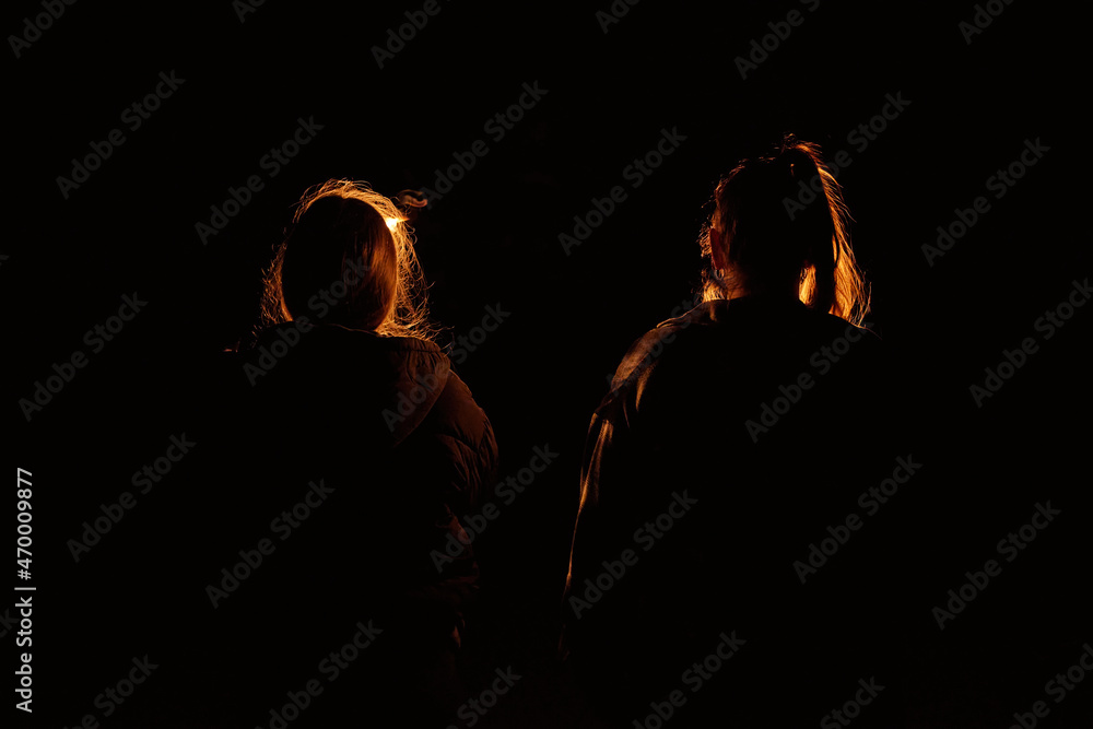Two unrecognizable women walk away from the viewer in a pitch black night holding a torch light which lets their hair glow