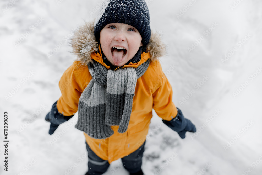 Charming little boy catches snowflakes with his tongue in a beautiful orange winter park. Cute baby playing in the snow. Winter entertainment for children. Top view Charming little boy catches snowflakes with his tongue in a beautiful orange winter park. Cute baby playing in the snow. Winter entertainment for children. Top view