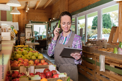 Portrait of happy female sales assistant in a grocery using digital tablet and talking on cell phone while taking inventory - Owner with digital tablet preparing online grocery order photo