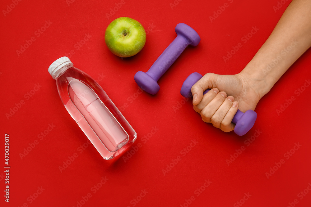 Female hand with dumbbells, apple and bottle of water on red background Female hand with dumbbells, apple and bottle of water on red background