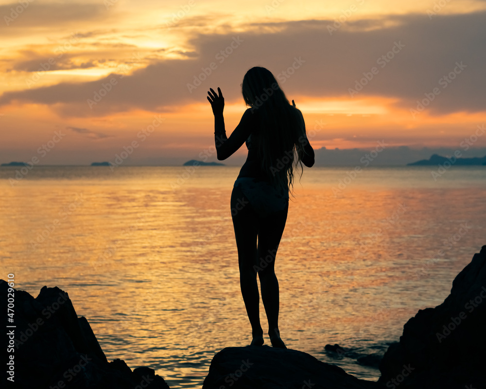 Silhouette of a young woman with a long hair standing by the sea and enjoying colorful sunset