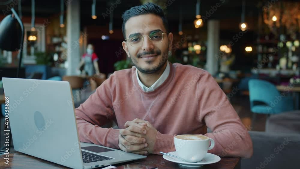 Slow motion portrait of handsome Middle Eastern guy in cafe sitting at table with laptop and coffee smiling looking at camera. People and lunch break concept. Slow motion portrait of handsome Middle Eastern guy in cafe sitting at table with laptop and coffee smiling looking at camera. People and lunch break concept.