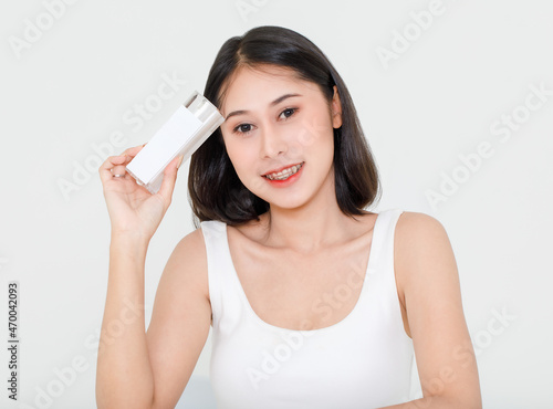 Portrait beauty shot of millennial young Asian short black hair female model in tank top vest undershirt holding natural serum facial cream skincare bottle package in hand on white background