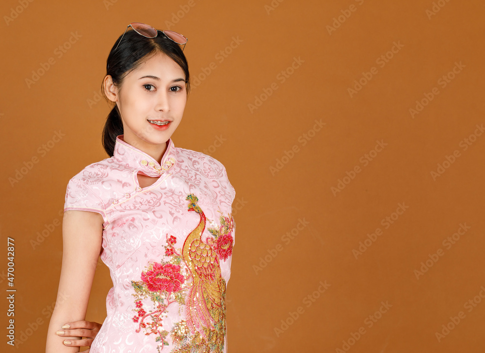 Portrait studio shot of millennial Asian female model in pink cheongsam qipao traditional festival peacock and flowers pattern dress and eyeglasses on head standing smiling posing on brown background Portrait studio shot of millennial Asian female model in pink cheongsam qipao traditional festival peacock and flowers pattern dress and eyeglasses on head standing smiling posing on brown background
