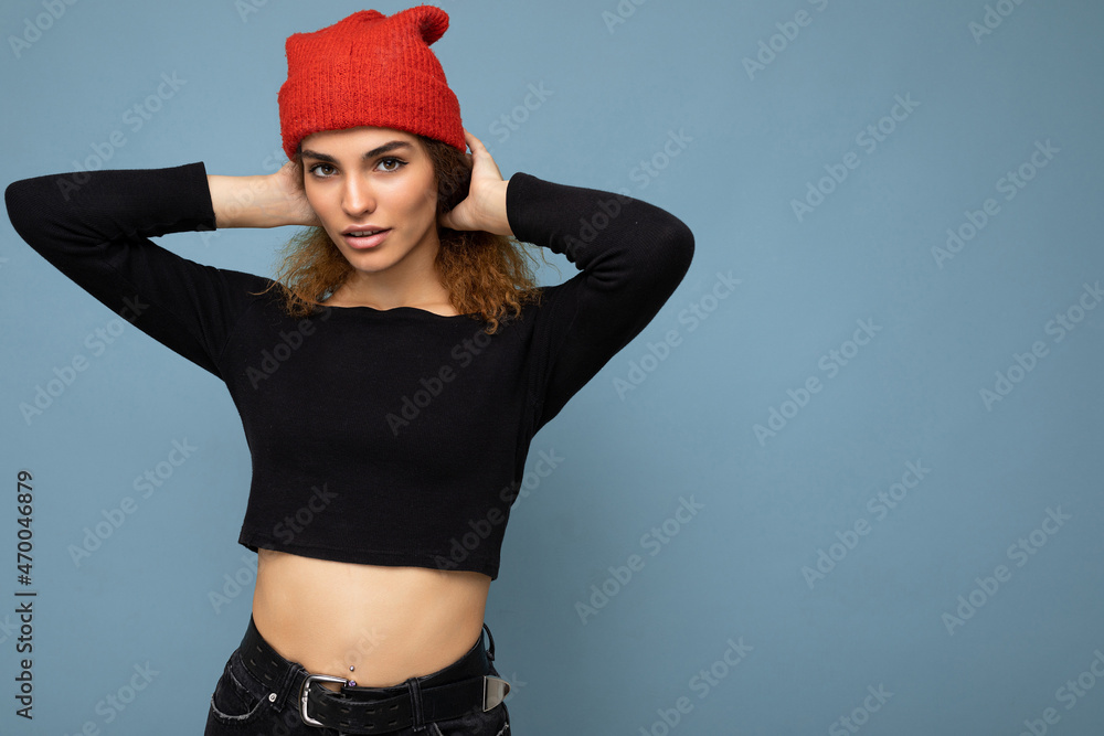 Photo of young positive cute nice brunette woman curly with sincere emotions wearing stylish black crop top and red hat isolated on blue background with copy space Photo of young positive cute nice brunette woman curly with sincere emotions wearing stylish black crop top and red hat isolated on blue background with copy space