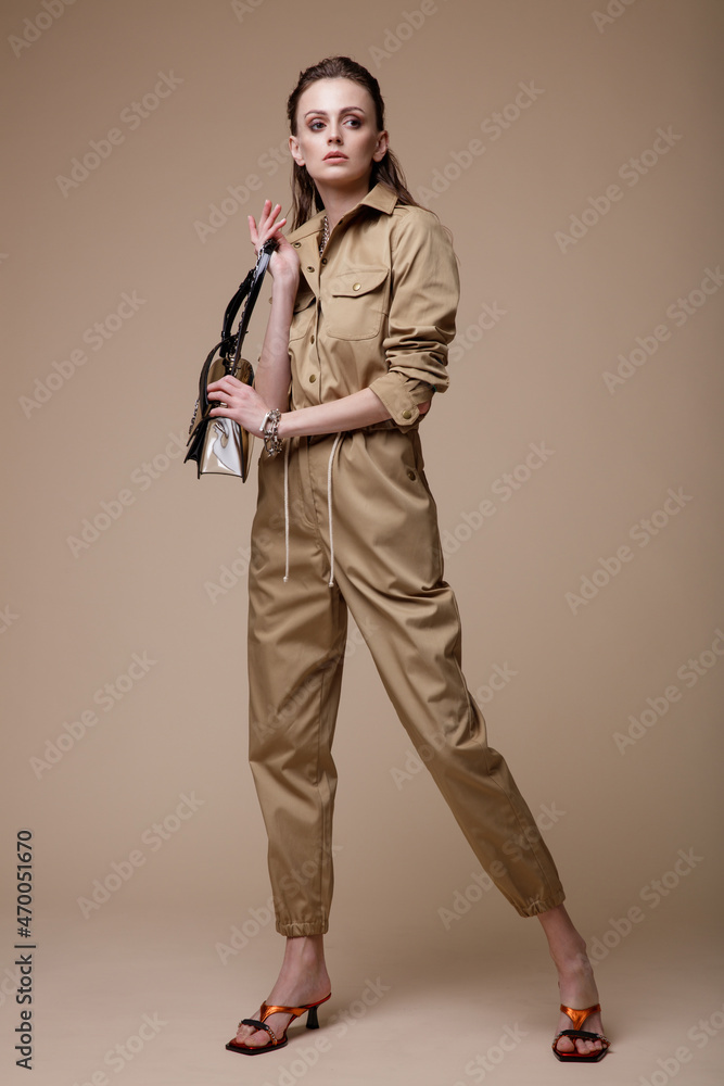Fototapeta premium High fashion photo of a beautiful elegant young woman in a pretty beige sand jumpsuit, accessories, handbag posing on brown background. Studio Shot. Slim figure. Make up, hairstyle
